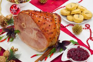 Roasted spiced ham on holiday dinning table, garnished with cloves, cinnamon sticks, hot chili pepper, and purple basil. Side dishes and Christmas ornaments around.